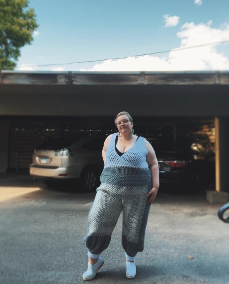 May include: A person wearing a light blue crocheted jumpsuit with a black bra peeking out. The jumpsuit has a textured, puffy design. The person is standing in a parking lot with a car in the background.