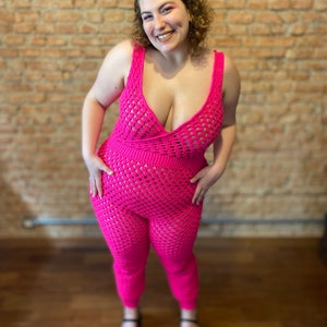 May include: A pink crocheted jumpsuit with a deep V-neckline and a fitted silhouette. The jumpsuit has a delicate open-weave pattern and is worn by a person standing in front of a brick wall.