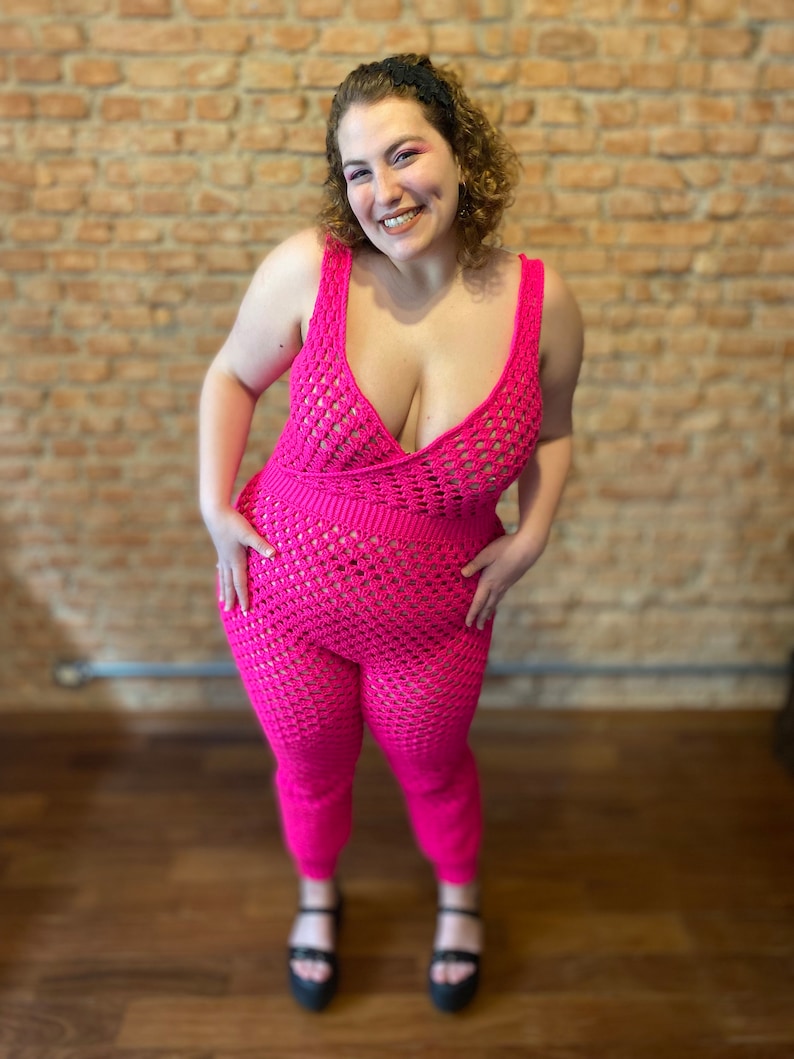 May include: A pink crocheted jumpsuit with a deep V-neckline and a fitted silhouette. The jumpsuit has a delicate open-weave pattern and is worn by a person standing in front of a brick wall.