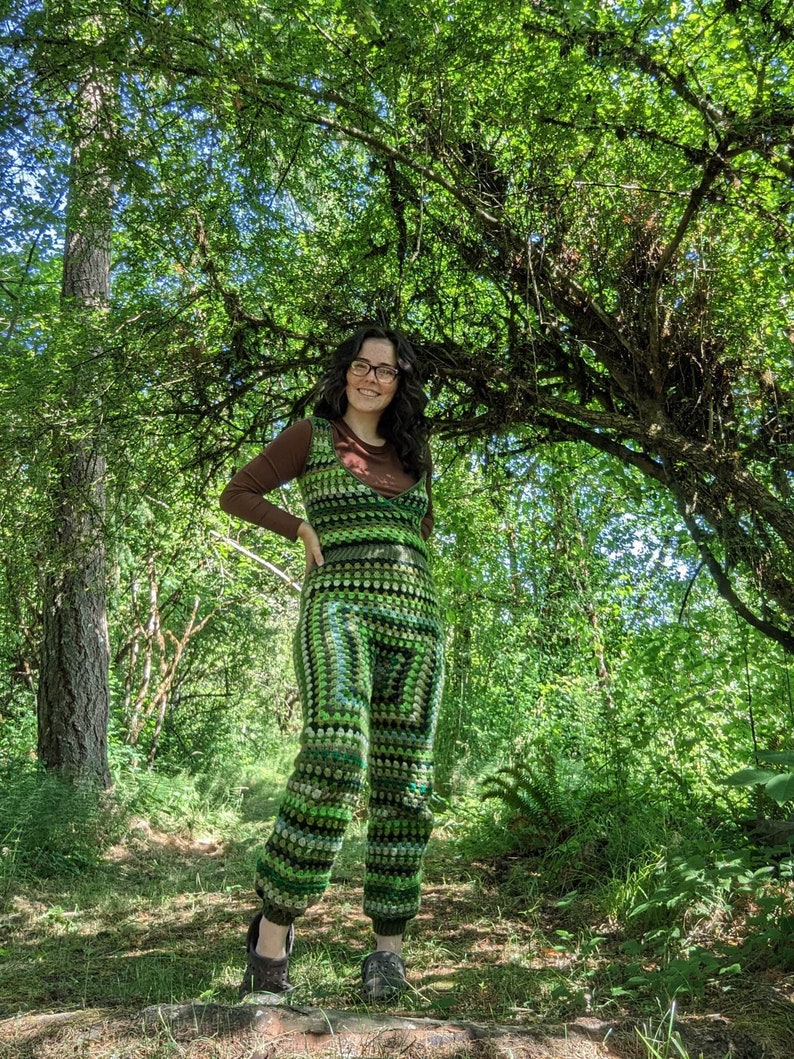 May include: A person wearing a green and brown crochet jumpsuit in a forest setting. The jumpsuit has a wide leg and a loose fit. The person is smiling and looking to the right.
