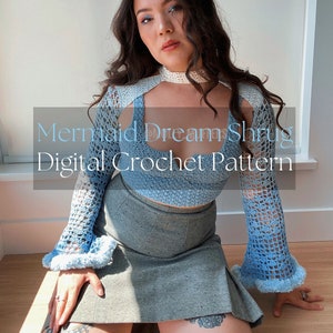 May include: A blue crochet shrug with a keyhole neckline and long sleeves. The shrug is worn over a white tank top and a gray skirt. The text "Mermaid Dream Shrug Digital Crochet Pattern" is superimposed on the image.