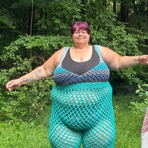 May include: A woman wearing a teal crochet top and matching pants. The top has a black under layer and a blue and gray crochet overlay. The woman has short purple hair and is standing in a grassy area.
