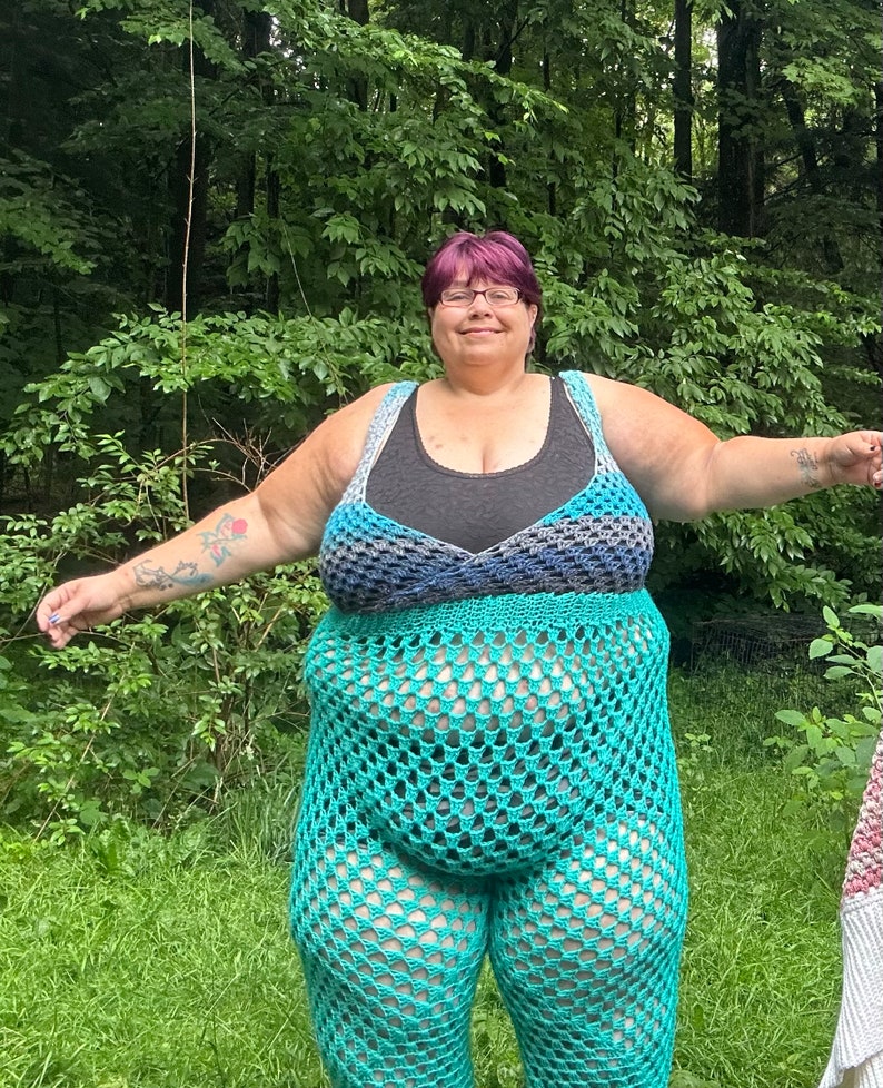May include: A woman wearing a teal crochet top and matching pants. The top has a black under layer and a blue and gray crochet overlay. The woman has short purple hair and is standing in a grassy area.