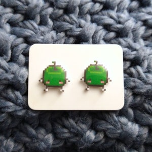 May include: A pair of green pixelated creature earrings on a white card. The creatures have small eyes and a single antenna.