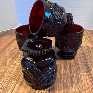 May include: Four ruby red vintage drinking glasses. Each glass has a detailed embossed pattern with a textured design. The glasses are arranged on a wooden surface, showcasing their intricate details and rich colour. A classic glassware set.