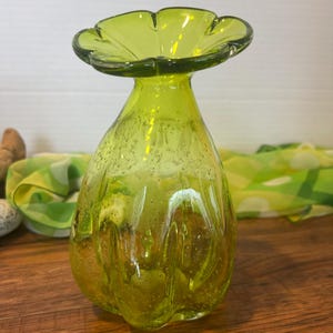 May include: A green glass vase with a wide, flared opening and a textured, bubbly surface. The vase has a pear-shaped body and a unique, ruffled rim.