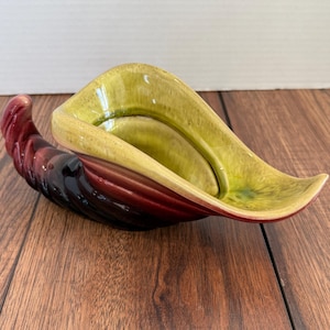May include: A ceramic, leaf-shaped dish with a glossy finish. The dish features a gradient of colours, transitioning from a deep burgundy to a light yellow-green. The design is a stylized, elongated form, suitable for decorative use.
