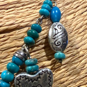 May include: Close-up of a turquoise beaded necklace with silver-toned heart and oval pendants. The heart pendant has a heart-shaped cutout. The oval pendant has an etched design. The necklace is on a woven background.