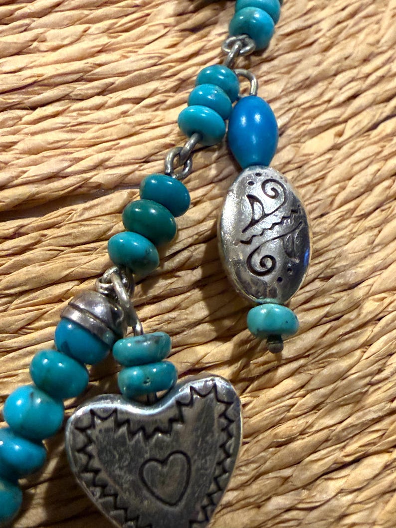 May include: Close-up of a turquoise beaded necklace with silver-toned heart and oval pendants. The heart pendant has a heart-shaped cutout. The oval pendant has an etched design. The necklace is on a woven background.