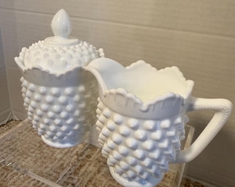 May include: A white hobnail creamer and covered sugar bowl set. The creamer has a handle and a ruffled rim. The sugar bowl has a lid with a knob. Both pieces have a textured, hobnail design.