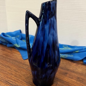 Op de afbeelding: Een blauwe en witte keramische vaas met een unieke vorm en een handvat. De vaas heeft een gemarmerde glazuur met witte strepen op een donkerblauwe achtergrond.