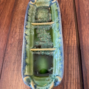 May include: A ceramic soap dish with a boat-like shape. The dish is glazed in shades of green and blue, with a textured, crystalline pattern in the center. The dish has two horizontal bars and a rounded base.