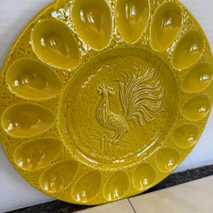 May include: Yellow ceramic egg serving platter with 12 egg-shaped indentations around the edge and a rooster relief in the center. The platter has a textured surface and a glossy finish, perfect for serving deviled eggs.