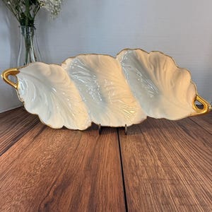 May include: A white, leaf-shaped serving dish with three compartments and gold trim. The dish is made of porcelain and has a glossy finish. The handles are also gold. The dish is displayed on a wooden surface.