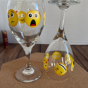 May include: Two clear wine glasses decorated with yellow emoji faces. One glass is upright, the other is upside down. The emoji faces have various expressions, including sad, surprised, and smiling. The glasses are on a brown surface.