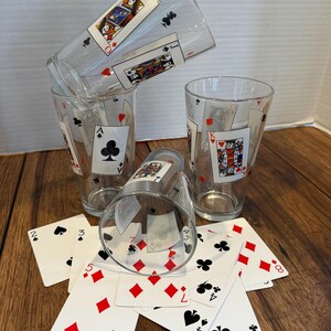 May include: Clear drinking glasses featuring playing card designs, such as the Ace of Clubs, King of Hearts, and Queen of Diamonds. Playing cards are scattered on a wooden surface near the glasses. A fun addition to any game night or poker evening.