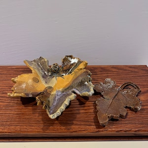 May include: Two ceramic leaf-shaped dishes, one in brown and one in a mix of brown, yellow, and white. The dishes are on a wooden surface.