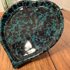 May include: Heart-shaped ceramic dish with a speckled teal and black pattern. The dish has a glossy finish and a raised edge. The dish is sitting on a wooden surface.