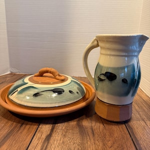 May include: A ceramic butter dish with a lid and a matching pitcher. The butter dish is round with a terracotta base and a lid with a handle. The pitcher has a handle and a decorative design with blue and black accents.