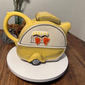May include: A yellow ceramic teapot shaped like a vintage camper trailer. The teapot features a white panel with strawberry accents, a curved handle, and a lid. The teapot is on a white circular base.