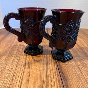 May include: Two ruby red glass mugs with ornate floral and geometric designs. Each mug has a pedestal base and a curved handle. The mugs are displayed on a wooden surface.