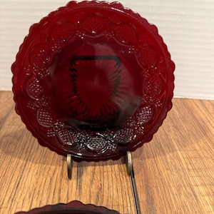 May include: A set of two ruby red glass plates with a scalloped edge and embossed fruit design. The plates are displayed on a gold-colored metal stand, showcasing their intricate details and rich color. The plates are a vintage style.