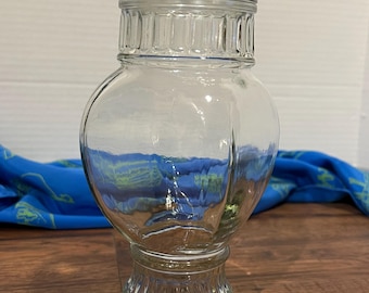 May include: A clear glass jar with a lid and a textured base. The jar is tall and has a rounded body.