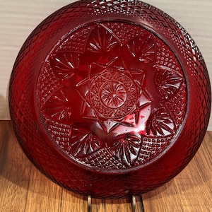 May include: A deep red glass plate featuring a detailed starburst and diamond pattern. The plate has a raised, textured design and is displayed on a metal stand. The plate is a decorative piece.