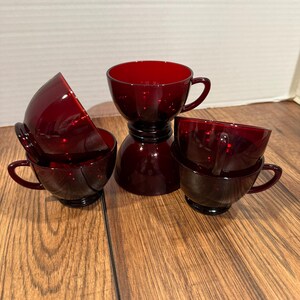 May include: A set of ruby red glass teacups and a matching creamer. The cups have a classic design with a rounded bowl and a small handle. The creamer has a unique stacked design. The set is displayed on a wooden surface.
