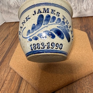 May include: A speckled beige ceramic crock featuring a blue hand-painted leaf design and the text FRANK JAMES TRIAH 1883-1990. The cylindrical vessel has a wide rim and a tapered base, with the blue lettering and graphic applied to the front surface.