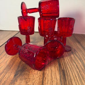 Puede incluir: Colección de copas de vino rojo rubí. Las copas están apiladas y dispersas, mostrando su diseño. Cada copa presenta un tallo y una copa con un patrón de racimo de uvas. Las copas están hechas de un material transparente.