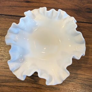 May include: A white, ruffled-edge bowl. The bowl has a glossy finish and a scalloped edge. The bowl is empty and sits on a wooden surface. The bowl's design is reminiscent of vintage or antique tableware.