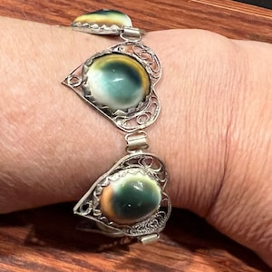 May include: A silver bracelet with three heart-shaped charms. Each charm features a round, iridescent shell with shades of green, brown, and yellow.