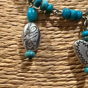May include: Close-up of a turquoise beaded necklace with silver-toned metal charms. The necklace features small, round turquoise beads and decorative oval and rectangular charms with etched designs. The background is a woven, textured surface.