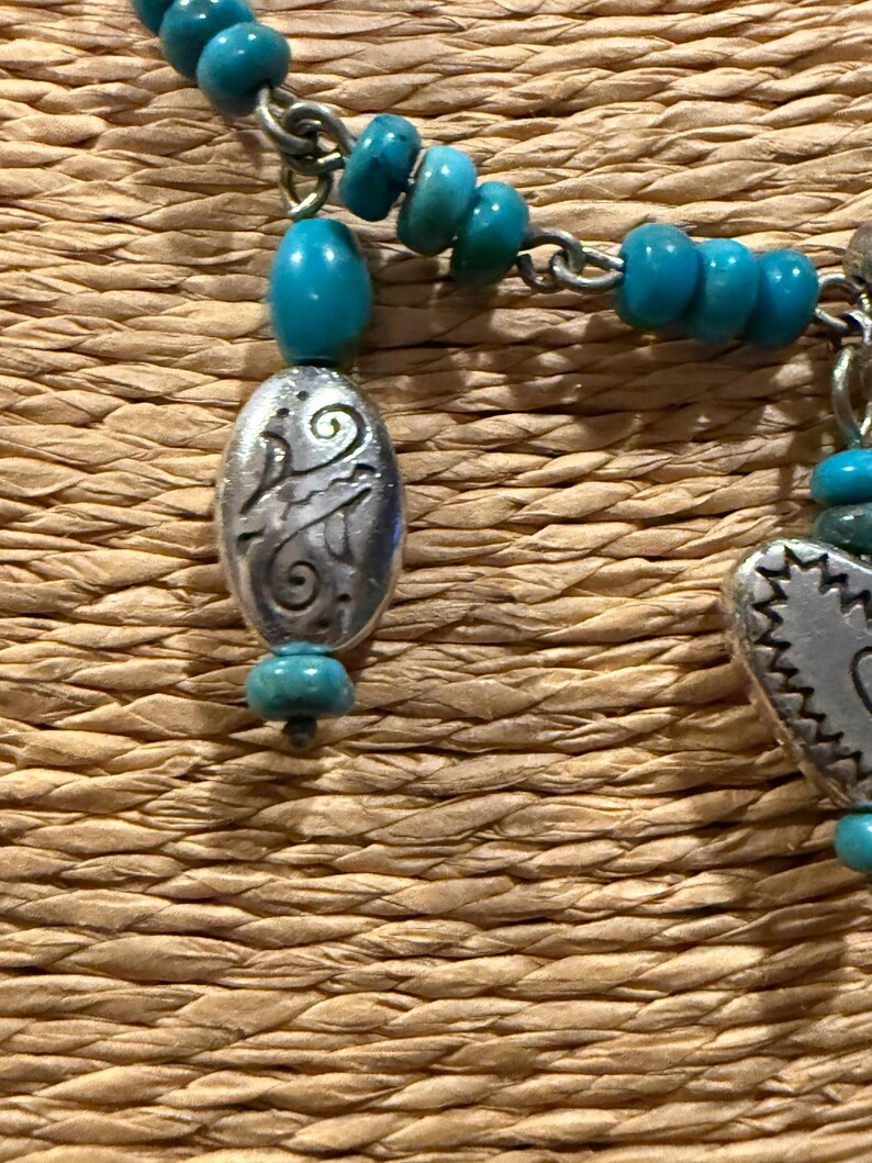 May include: Close-up of a turquoise beaded necklace with silver-toned metal charms. The necklace features small, round turquoise beads and decorative oval and rectangular charms with etched designs. The background is a woven, textured surface.