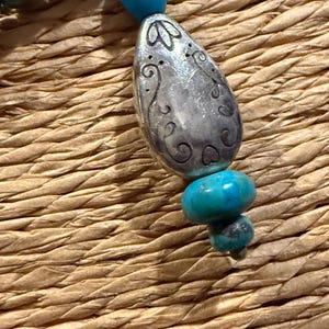 May include: Close-up of a turquoise beaded necklace with a silver pendant. The pendant is an elongated oval shape with etched floral designs. The necklace features various sizes of turquoise beads.