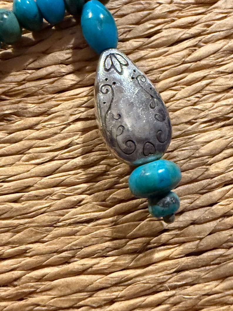 May include: Close-up of a turquoise beaded necklace with a silver pendant. The pendant is an elongated oval shape with etched floral designs. The necklace features various sizes of turquoise beads.