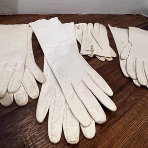 Op de afbeelding: Een set van vijf witte leren handschoenen. De handschoenen zijn allemaal lang en hebben een gladde textuur.