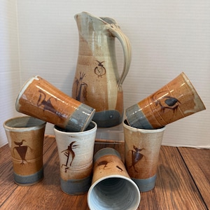 May include: A handmade ceramic pitcher and six matching tumblers. The pitcher and tumblers are tan with brown and gray accents. Each tumbler features a unique brown illustration. The set is displayed on a wooden surface.