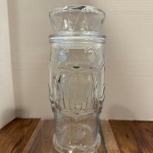 May include: A clear glass jar with a lid, featuring a decorative design of a bear. The jar has a cylindrical body and a rounded lid. The bear design is etched into the glass, adding a vintage touch. The jar is displayed on a clear acrylic stand.
