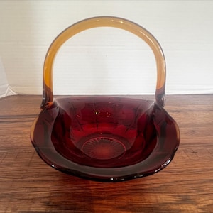 May include: A vintage ruby red glass basket with a clear amber handle. The basket has a round, shallow bowl shape with a decorative pattern. The handle arches gracefully above the bowl, creating a classic design.