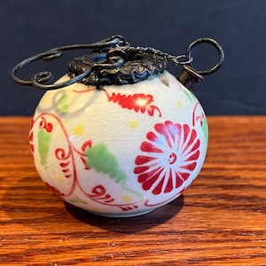 Peut inclure: Une petite théière en céramique blanche avec un motif floral rouge et vert. La théière a un couvercle en métal avec une poignée décorative et un bec.