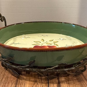 May include: Oval serving dish with a green exterior and a cream-colored interior featuring red apples and the word "Apples". The dish has a brown rim and is supported by a decorative metal stand with handles.