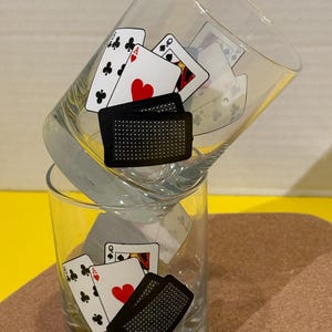 May include: Two clear drinking glasses with playing card designs. One glass is stacked on top of the other. The playing card designs include the Ace of Hearts, Queen of Diamonds, and the 8 of Clubs.