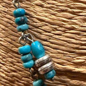 May include: Close-up of a turquoise beaded necklace with silver accents. The necklace features round and oval turquoise beads strung on a silver chain. The background is a woven, textured surface.