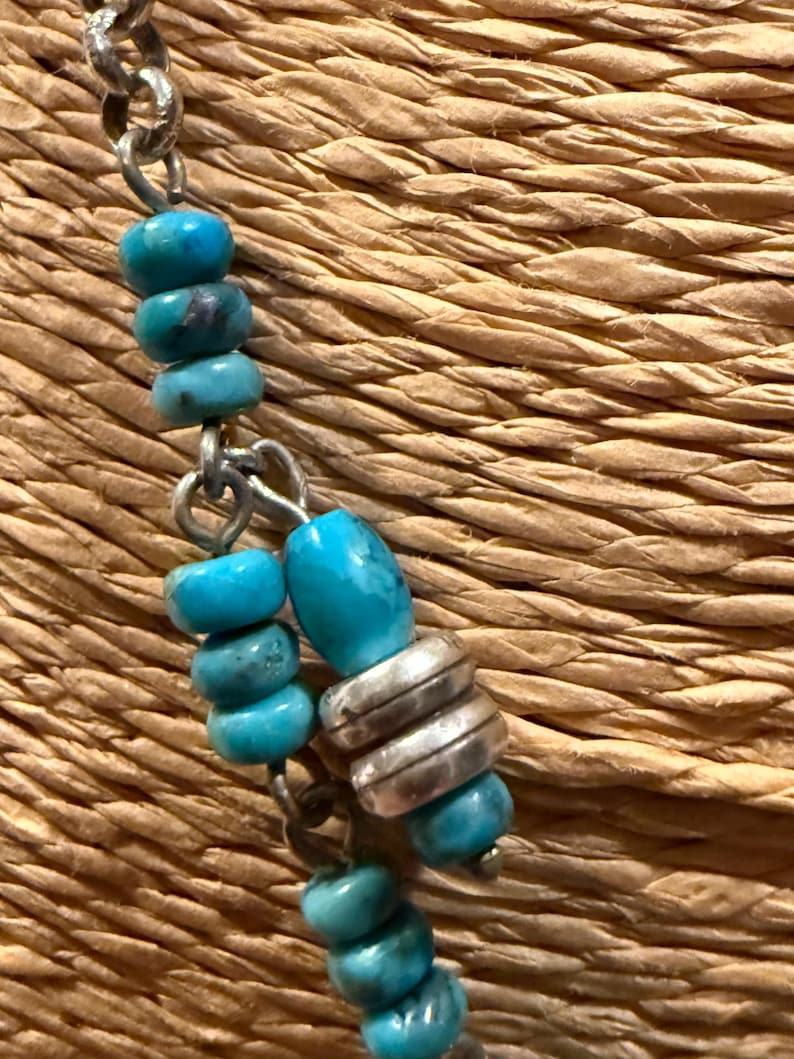 May include: Close-up of a turquoise beaded necklace with silver accents. The necklace features round and oval turquoise beads strung on a silver chain. The background is a woven, textured surface.