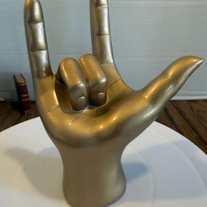 May include: A gold-colored hand sculpture in the "I love you" sign. The sculpture is on a white surface, with a wooden table and a small book in the background. The hand is in a gesture with the thumb, index, and pinky fingers extended.
