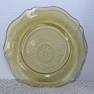 May include: A vintage yellow-tinted glass plate with a scalloped edge. The plate features a detailed embossed floral and geometric pattern around the rim and center. The plate is likely from the Depression era and is a collectible item.