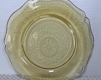 May include: A vintage yellow-tinted glass plate with a scalloped edge. The plate features a detailed embossed floral and geometric pattern around the rim and center. The plate is likely from the Depression era and is a collectible item.