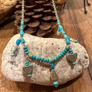 May include: A turquoise beaded necklace with silver-toned heart and teardrop charms. The necklace is displayed on a light-colored rock against a wooden background. The necklace has a chain and turquoise beads.
